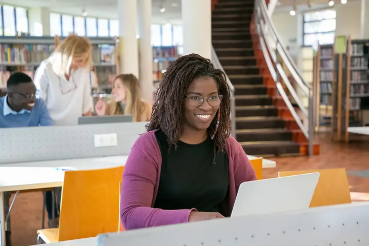 Estudante de óculos sorrindo, com o computador aberto na mesa da biblioteca