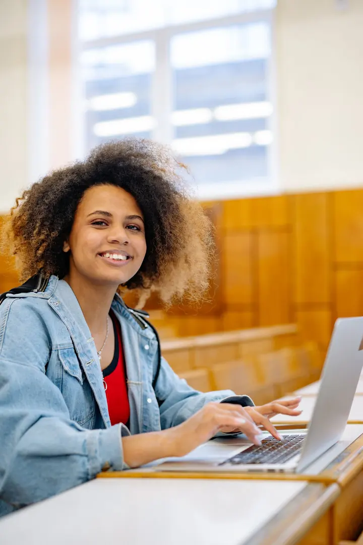 Estudante sorrindo em frente ao computador aberto, numa sala de aula com bancadas de madeira